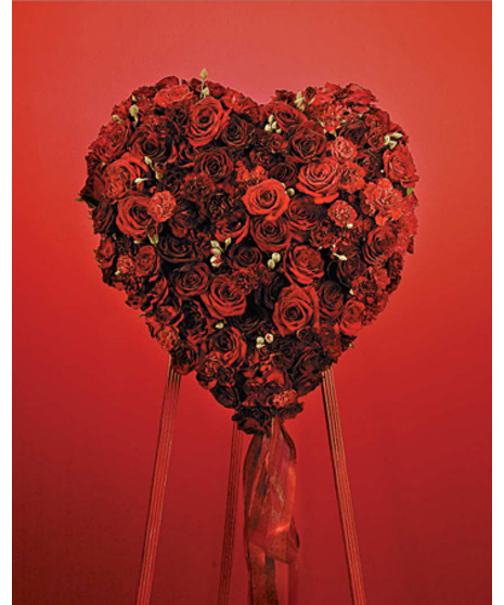 The solid heart is covered in red and burgundy carnations, miniature carnations, roses, and spray roses with red ribbon and myrtle comes on green wire easel stand 