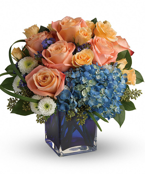 cobalt blue cube with blue hydrangea, peach roses and spray roses, white buttons, purple statice, lily grass, seeded eucalyptus, salal to create low compact round arrangement