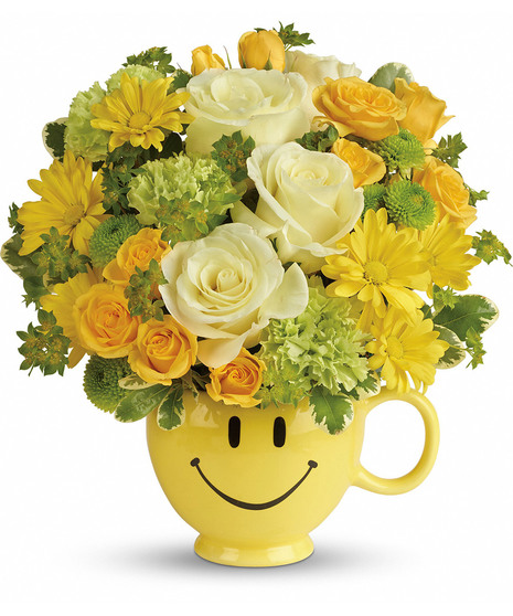 ceramic mug with happy face emoji has daisy, roses, spray roses, mums, carnations, buttons nestled in  pittosporum. The mug has a handle and is food safe