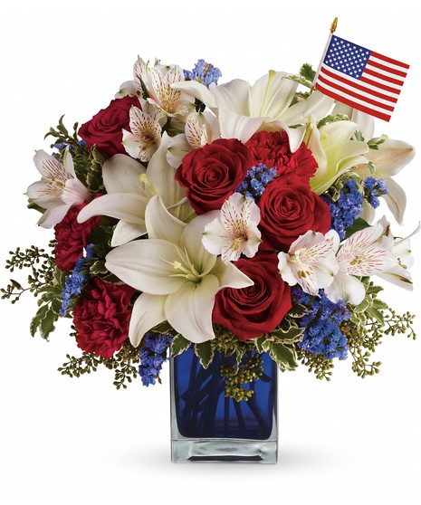 Red roses, carnations, white lilies and alstroemeria, blue statice and an American flag fill blue glass cube. 