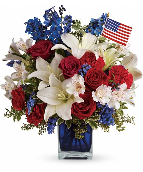 Red roses, carnations, white lilies and alstroemeria, blue statice and an American flag fill blue glass cube. 