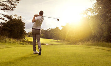 A golfer finishes a swing on a well-maintained course, illuminated by a setting sun against a backdrop of trees.
