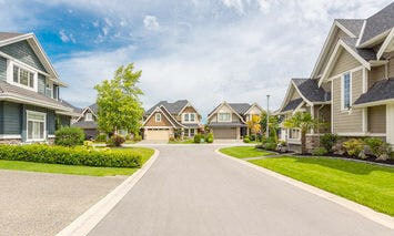 Suburban street with several modern houses lined along a wide, clean road; front yards are well-maintained with grass and trees, under a partly cloudy sky.