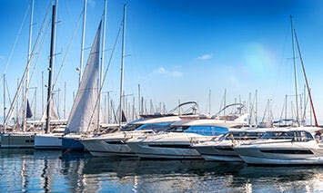 Yachts are docked in a marina, their sleek designs and masts reflecting in the calm water, against a clear blue sky backdrop.