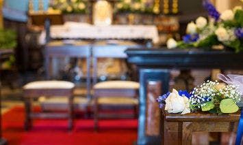 Flower arrangements rest on a wooden table, set in a church decorated for a wedding or ceremony with chairs, an altar, and red carpet in the background.