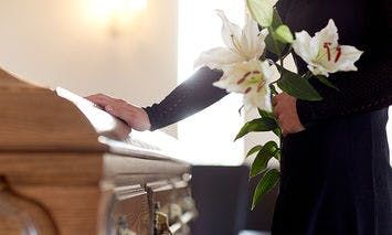 A person dressed in black, holding white lilies, places a hand on a wooden casket in a softly lit room, suggesting a funeral or memorial service.
