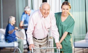 An elderly man using a walker receives assistance from a smiling caregiver in green scrubs in an outdoor care facility, with other seniors and a caregiver in the background.