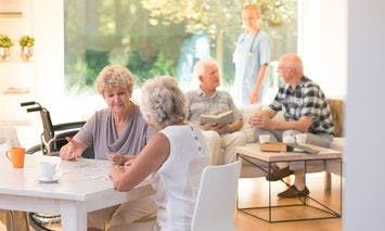 Elderly individuals sit and interact at a table and on a couch, in a well-lit room with large windows; a caregiver stands in the background. The environment appears calm and communal.