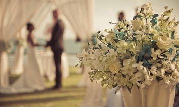 A bouquet of white and light blue flowers in a vase stands in the foreground, while a bride and groom hold hands in the blurred background beneath an elegant outdoor canopy.